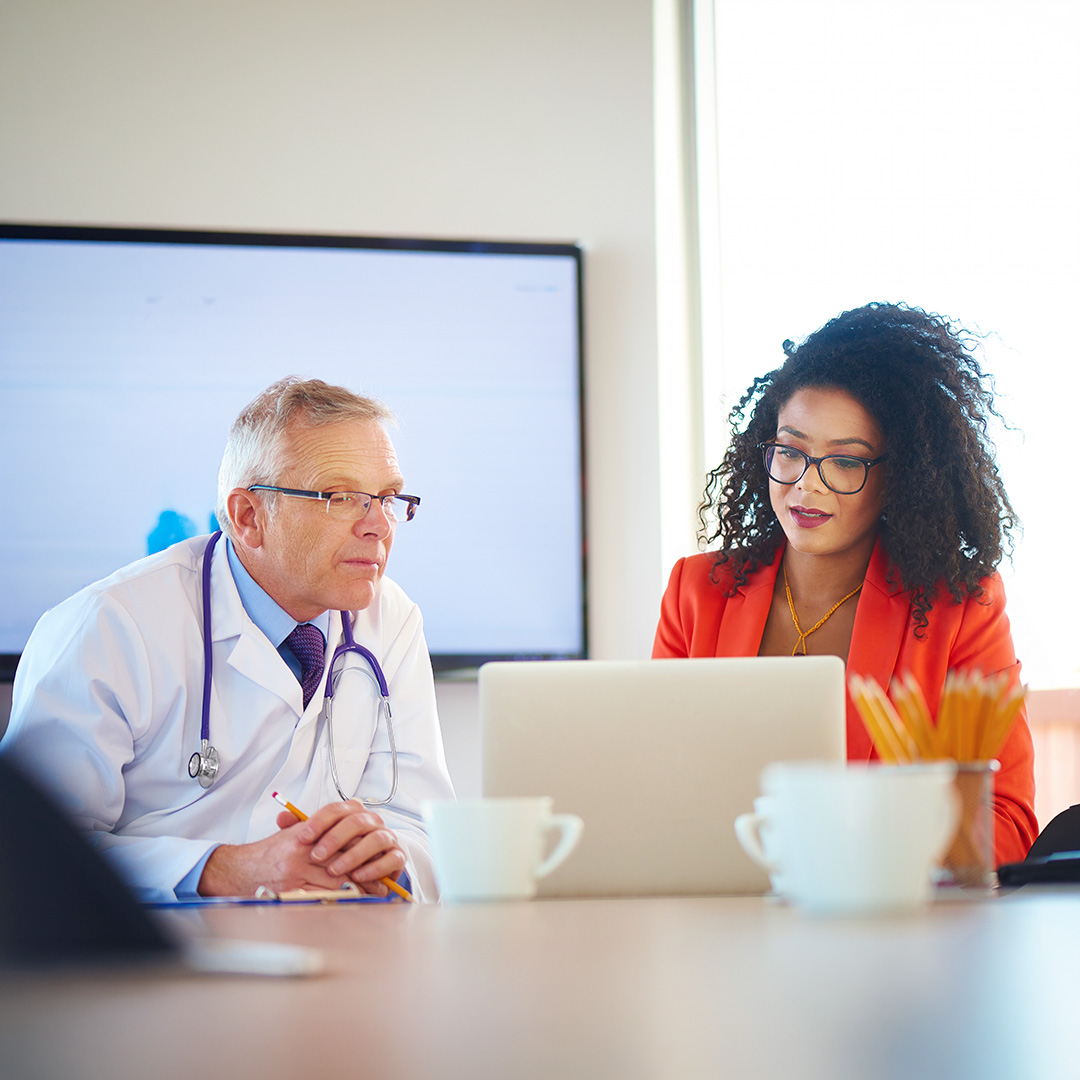 Doctor in white coat with stethoscope and woman in orange blazer reviewing laptop together at conference table.