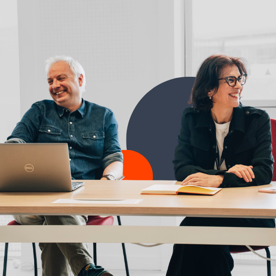 Deux professionnels souriant à une table de conférence avec un ordinateur portable et des documents dans un cadre de bureau moderne et lumineux.