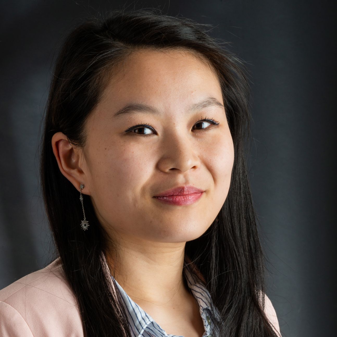 Close-up professional portrait of a young woman with long dark hair, wearing a light jacket and patterned shirt, and a dangling earring, photographed against a dark, softly lit background in a studio setting.