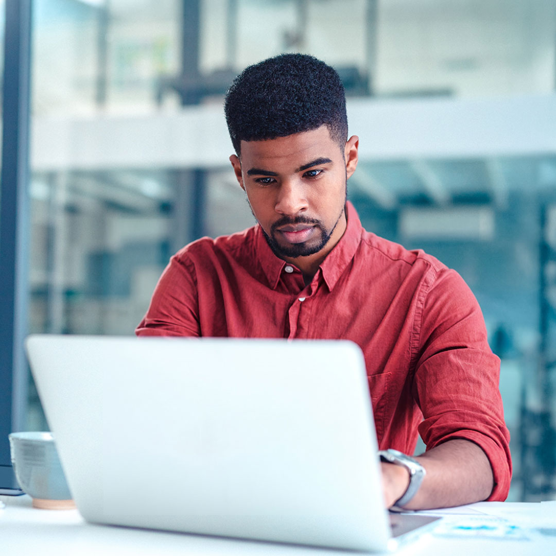 Young man in red shirt working intently on laptop in modern office with coffee cup nearby and blurred background.