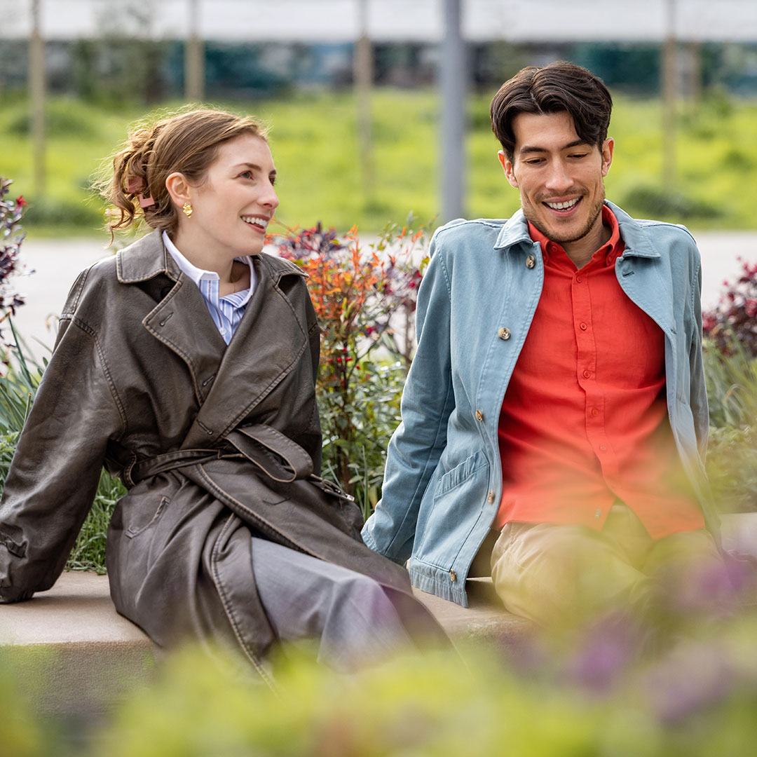 A smiling woman in a brown jacket and man in a red shirt and blue jacket sit together outdoors in a garden setting.