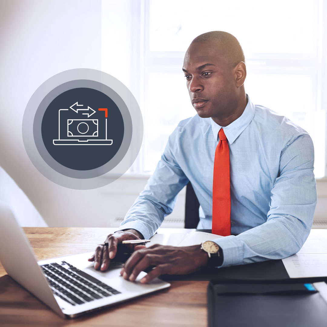 Professional man in light blue shirt and orange tie working on laptop at wooden desk in bright office setting.