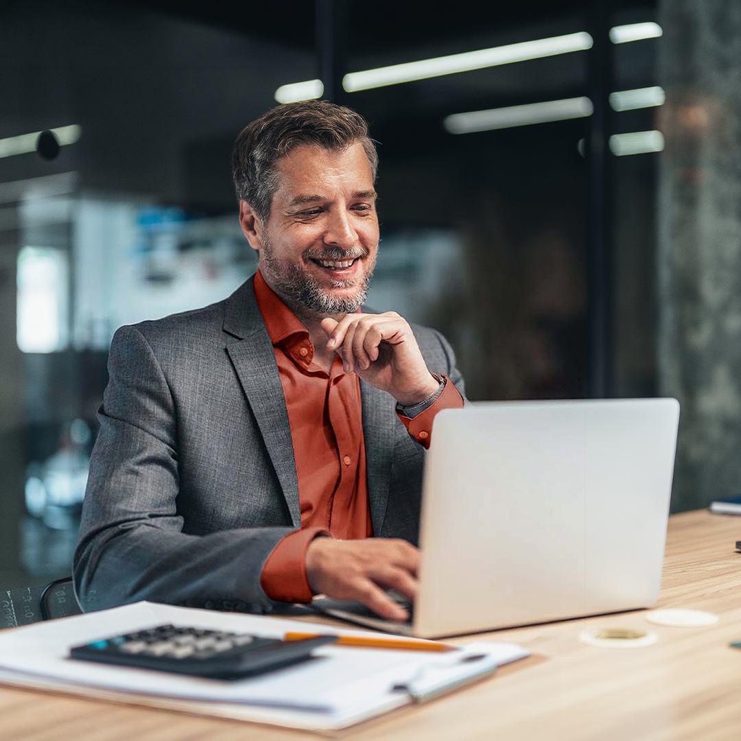 Smiling businessman in gray suit and orange shirt working on laptop at modern office desk with calculator and notebook.