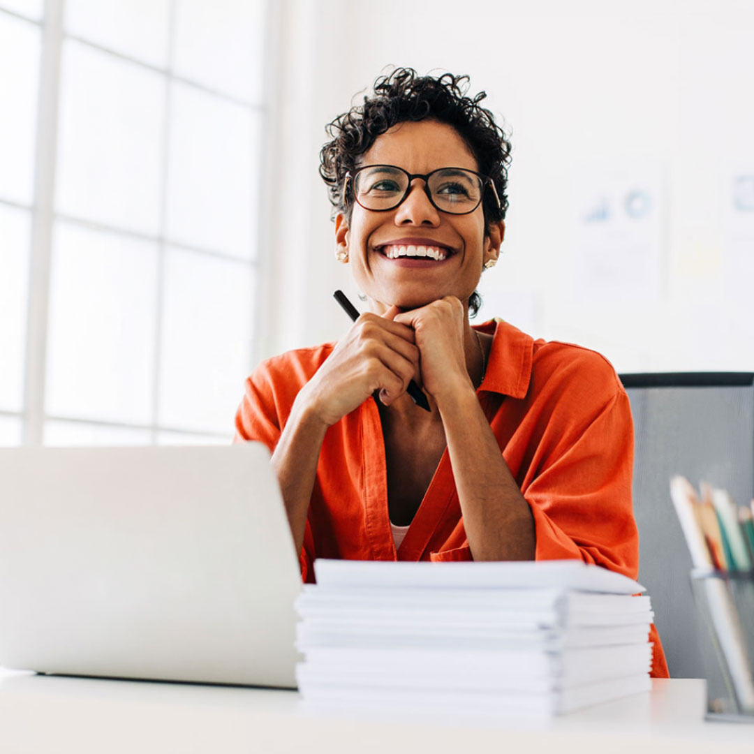 Smiling woman in orange blazer and glasses holding pen, looking up thoughtfully at desk with laptop and envelopes.