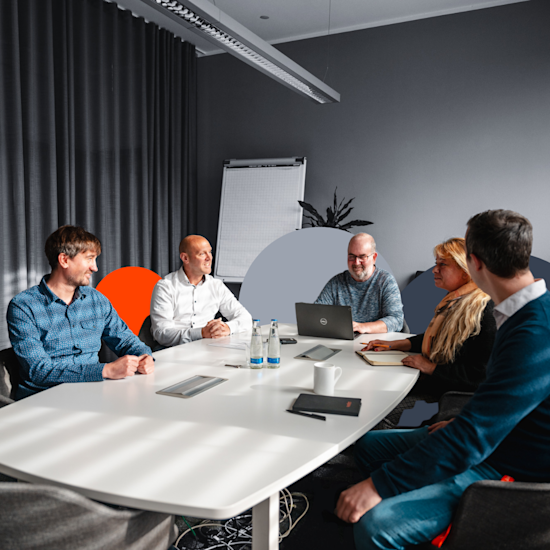 Business team meeting in modern gray conference room with white table, laptop, and orange accent chair.