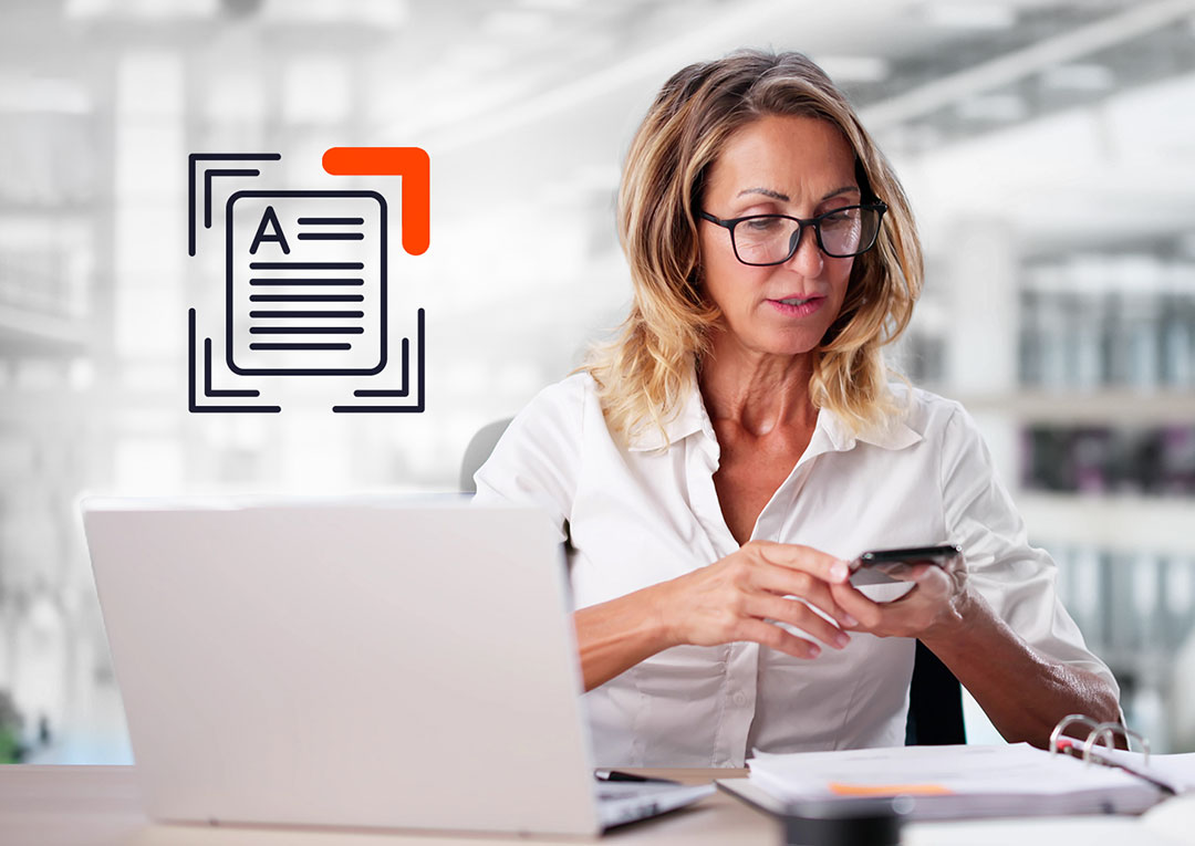 Woman in glasses scanning documents with her smartphone at desk with laptop, document icon overlay in upper left corner of image.