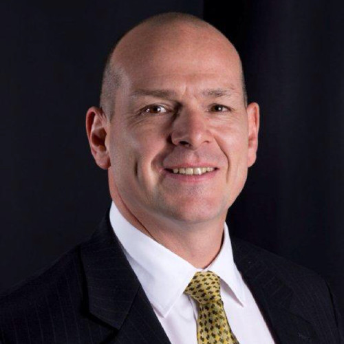 Professional headshot of a man with a shaved head, wearing a dark suit, white shirt, and patterned gold tie. The subject faces the camera with a slight smile against a dark, studio-style background.