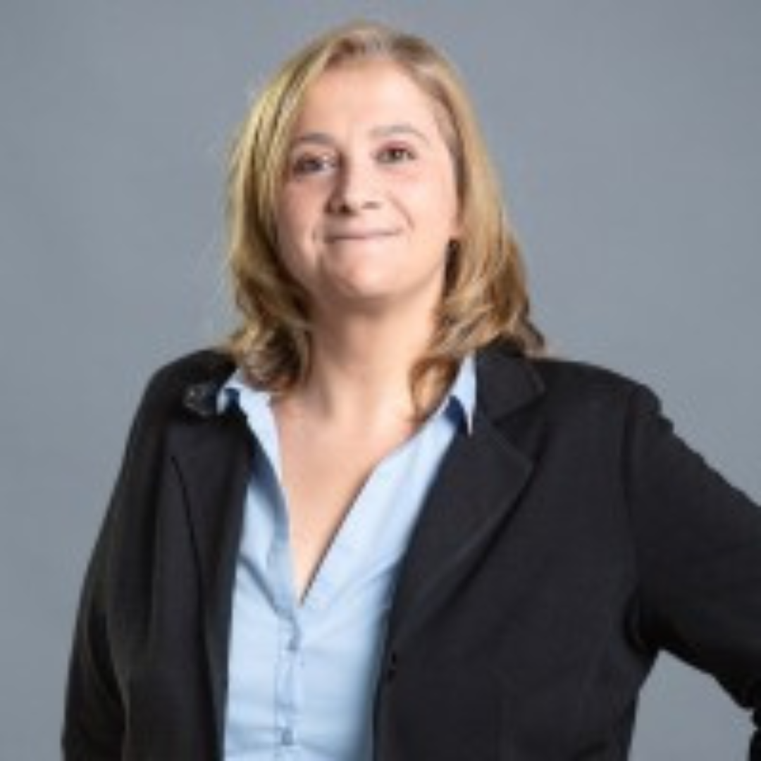a blond woman wearing a dark blazer and light blue shirt posing against a gray background