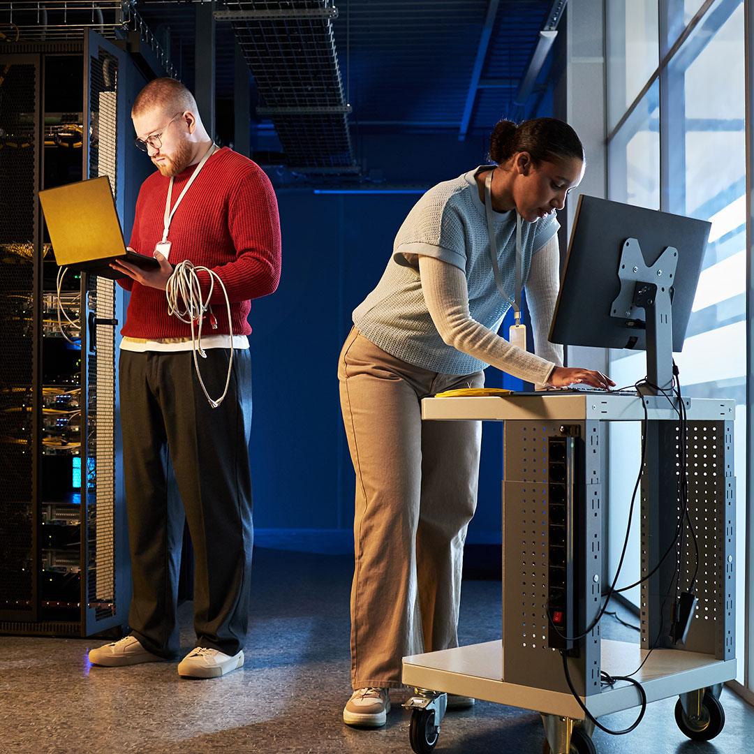 Two IT professionals working in a server room, one holding a laptop near server racks, the other at a standing desk.