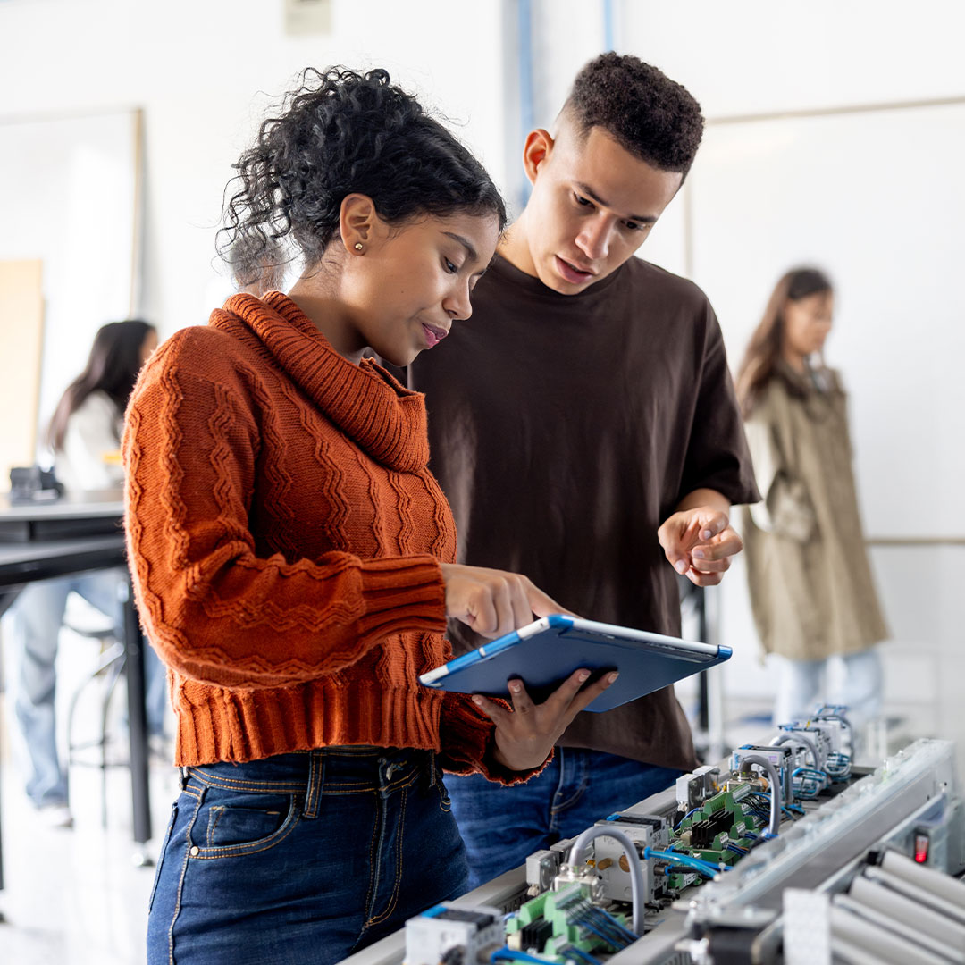 Two students in orange and brown sweaters examining a blue tablet while working with electronic circuit boards.