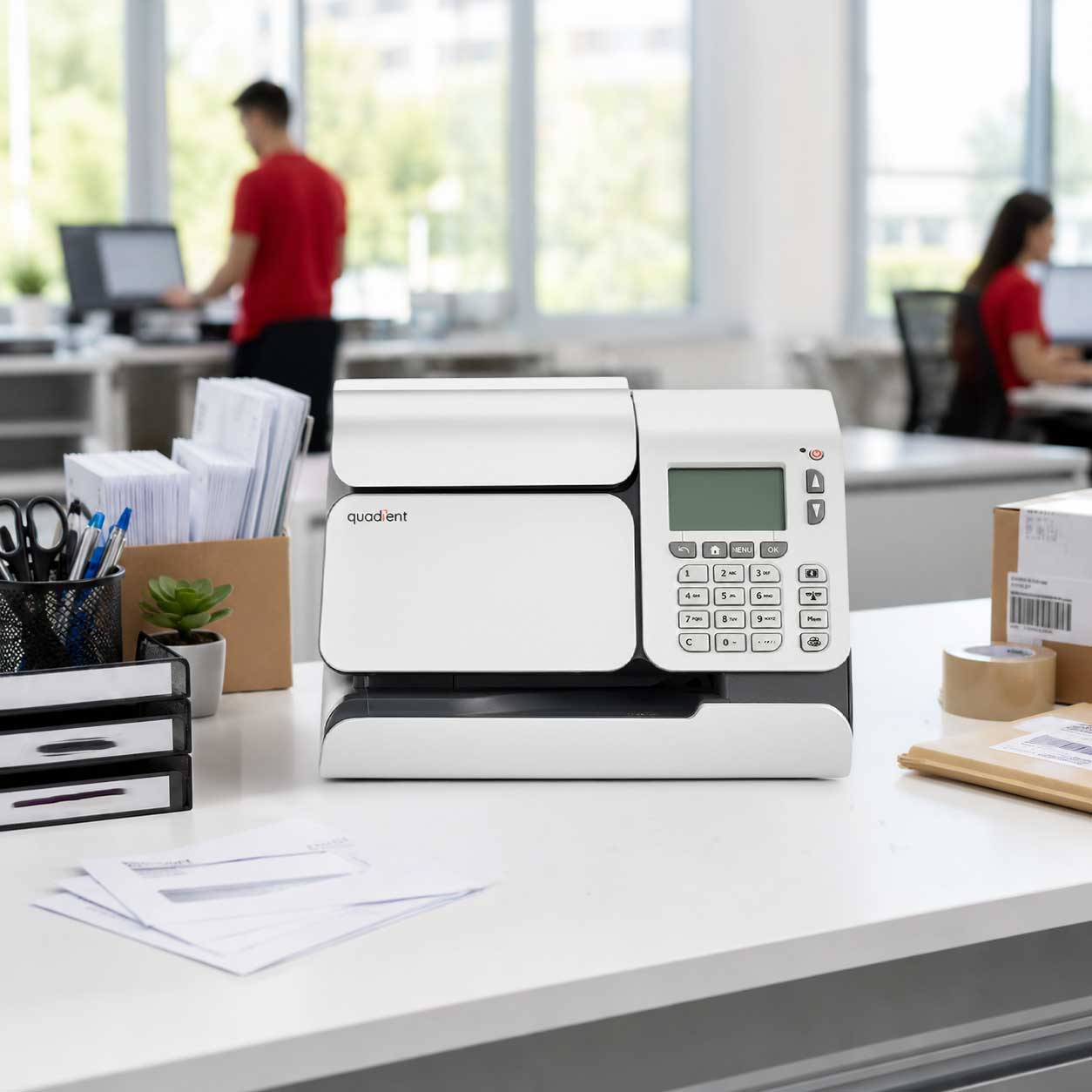 Quadient iS-280c franking machine on office desk with stationery, envelopes, and employees in red shirts in background.