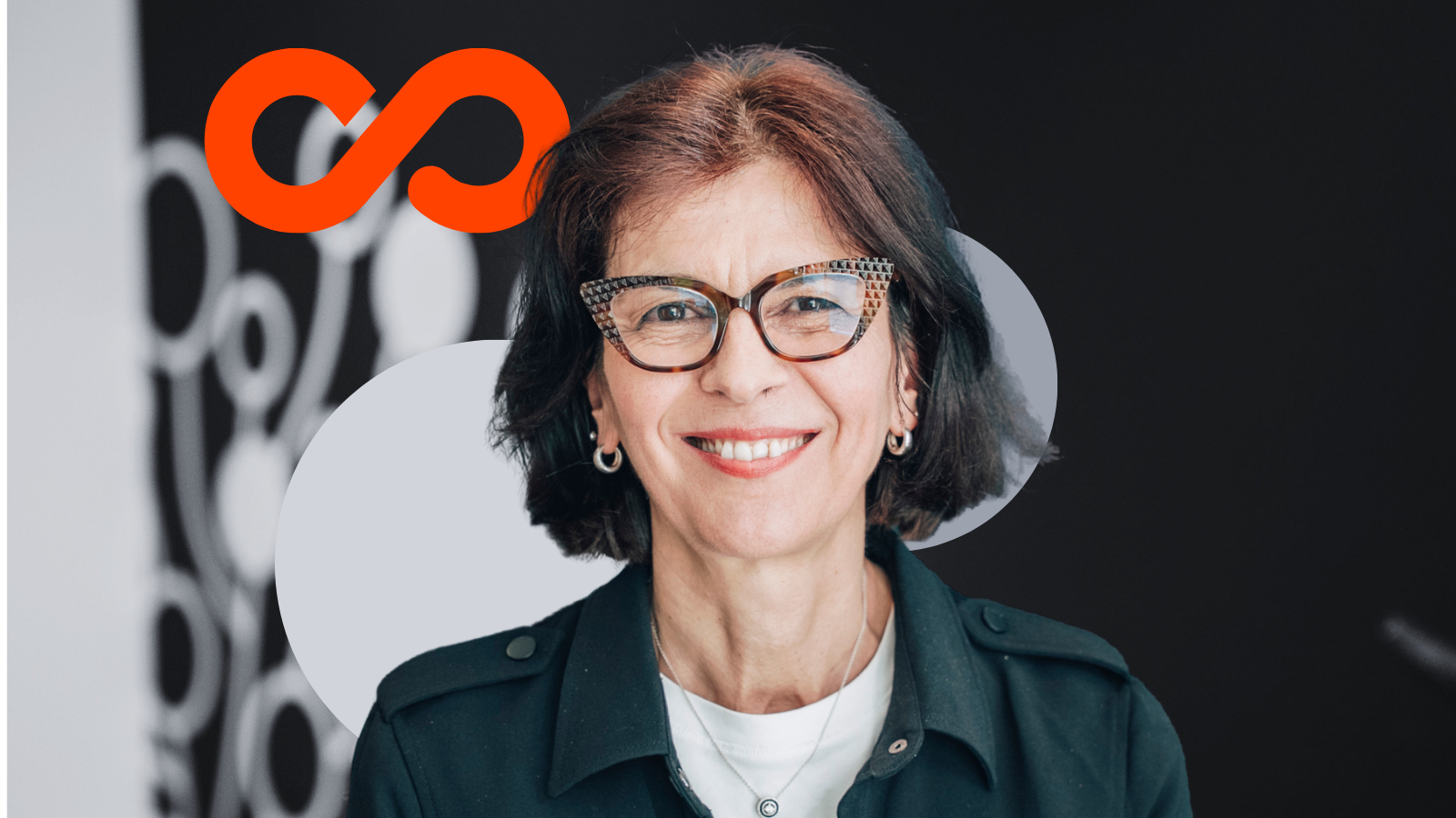 Smiling woman with short dark hair wearing patterned eyeglasses and dark green jacket against backdrop with orange infinity logo.