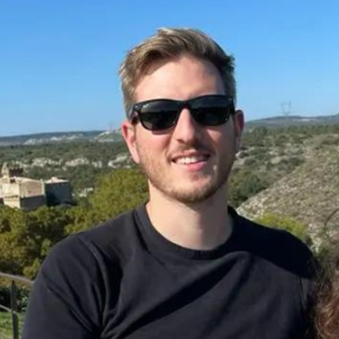 Un homme blond portant des lunettes de soleil et un chandail noir dehors avec des collines, des arbres et un ciel bleu en arrière-plan.