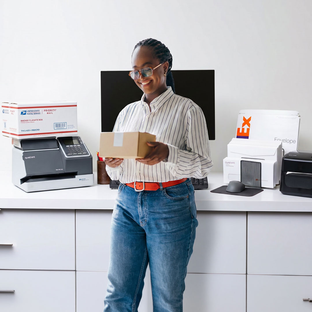 Woman in striped shirt and glasses smiling while holding a cardboard box, standing at desk with shipping equipment.
