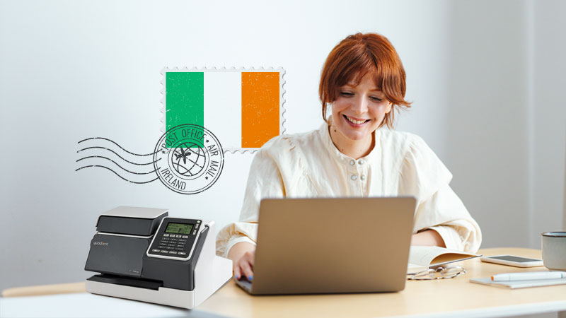 Woman with red hair smiling at laptop on desk, Irish flag postmark graphic on wall, franking machine beside her.
