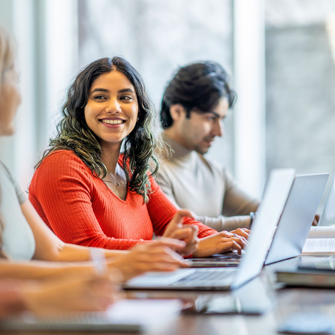 Smiling woman in orange top working on laptop in bright office with colleagues in background