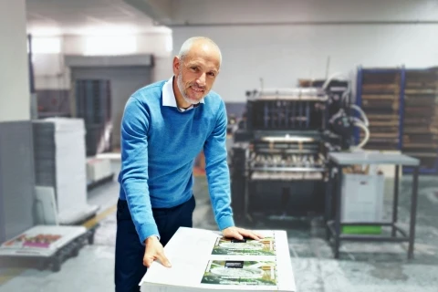 Smiling man in blue sweater examining printed sheets at a table in a printing facility with industrial equipment behind him.