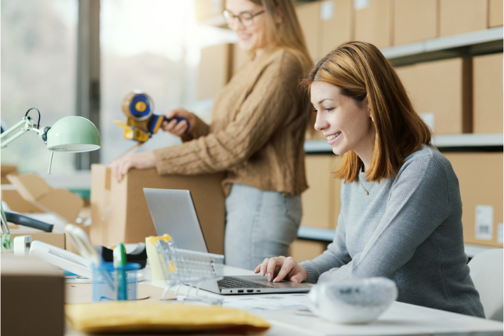 Two office employees prepare outgoing mail in a shipping workspace. One person works on a laptop while another seals a cardboard box in the background, with packing supplies and mailroom materials visible.