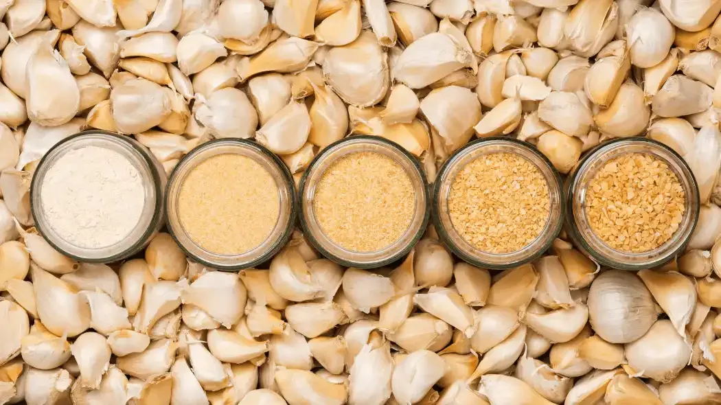 Various glass bottles filled with garlic powder behind backdrop of garlic cloves
