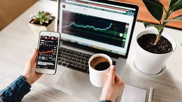 Person sitting in front of a laptop holding phone in left hand and coffee mug in the right hand