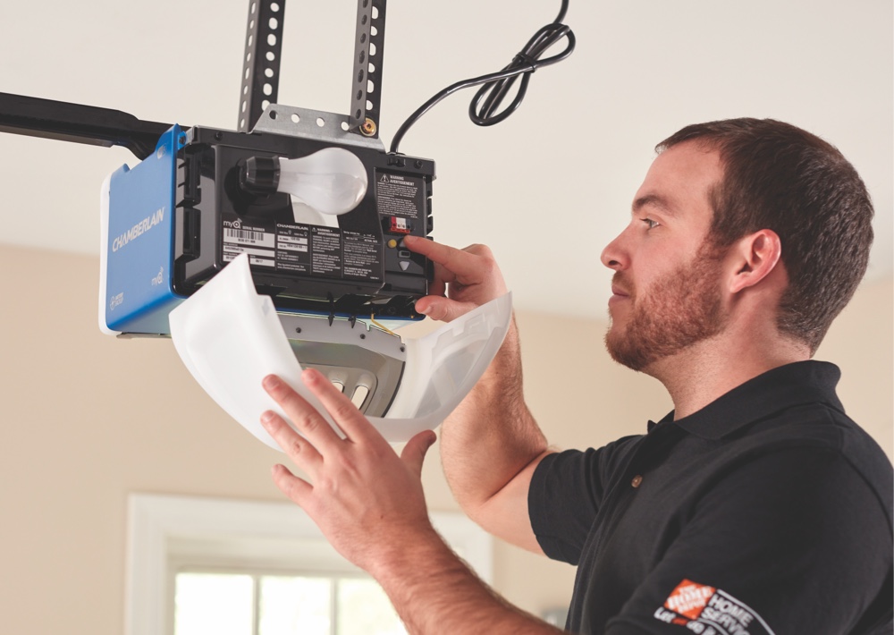 A technician wearing a black Home Depot shirt installs or repairs a Chamberlain garage door opener. He is adjusting internal components of the ceiling-mounted unit, with the light cover open and a power cord hanging above. The scene takes place indoors with daylight coming through a nearby window.