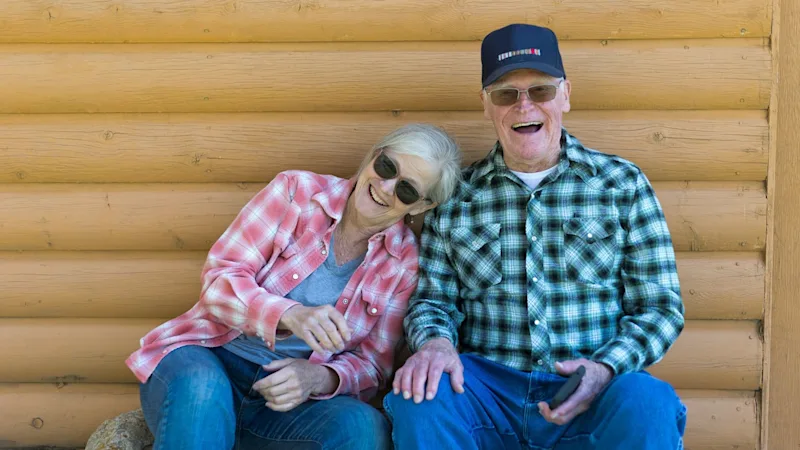 An older couple laughing together on a bench outside a log cabin, relaxed about their estate planning
