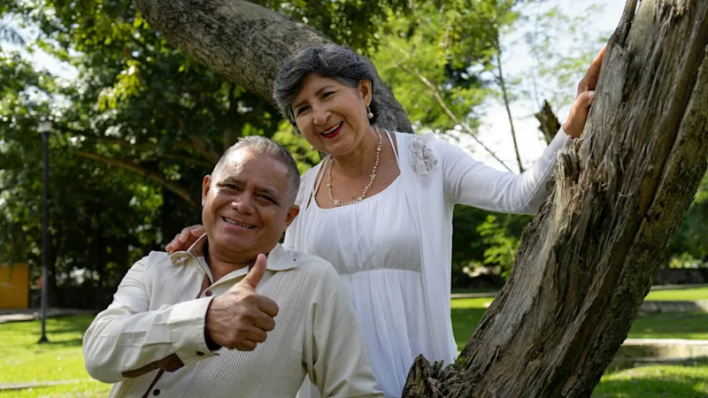 A happy older couple standing outdoors by a tree, smiling and giving a thumbs up, looking healthy and secure.