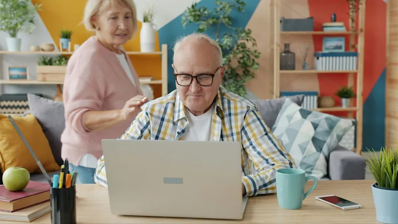 A couple in their living room, with the man sitting at a laptop to create an LPA online while his wife stands supportively behind him.