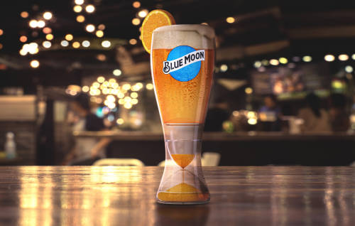 Interior of a bar with a Blue Moon branded glass on a table with Blue Moon beer in it and an orange wedge on the rim