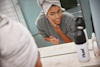 woman looking in the mirror washing her face with her hair in a towel and a huel shaker on the side