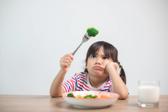 a child frowning at a piece of broccili on a fork with a plate of veg in front of them
