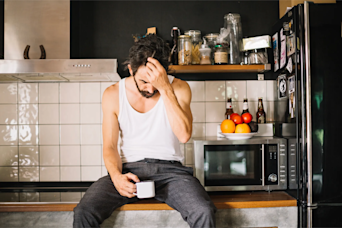 man sat on kitchen counter with his head in his hand and holding a coffee
