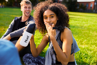 woman sat outside holding a huel shaker looking at the camera smiling and covering her mouth