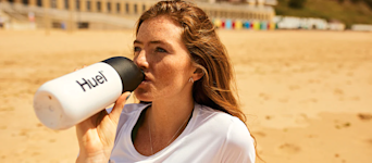 Woman on a beach in the sun drinking from a Huel shaker