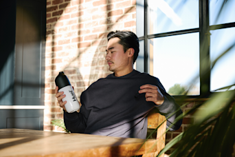 man sat on a chair at a table by a window holding a huel shaker with the sun shining through