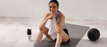 Woman sitting on a black yoga mat in a grey fitness studio, next to a huel shaker and a black medicine ball.