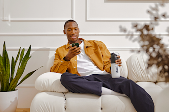 Man on a white couch, on his phone, holding a Huel shaker