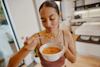 Woman eating Huel Instant Hot Meal from a bowl with a spoon while stood in the kitchen