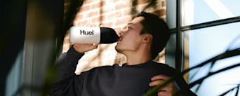 Image of a man reclining in a sunlit room on a chair while drinking from a black and white Huel shaker bottle