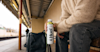 person sat on a bench at a train station holding a Huel Banana Ready-to-drink bottle with a bag next to them on the ground