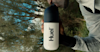 Huel shaker in the centre with a man holding and drinking from it while outside with trees in the background