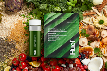 Huel Daily Greens pouch and bottle laying on a bed of different fruits and vegetables