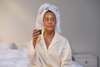 woman sat on a bed with a towel on her head holding a glass of Huel Daily Greends Powder Drink