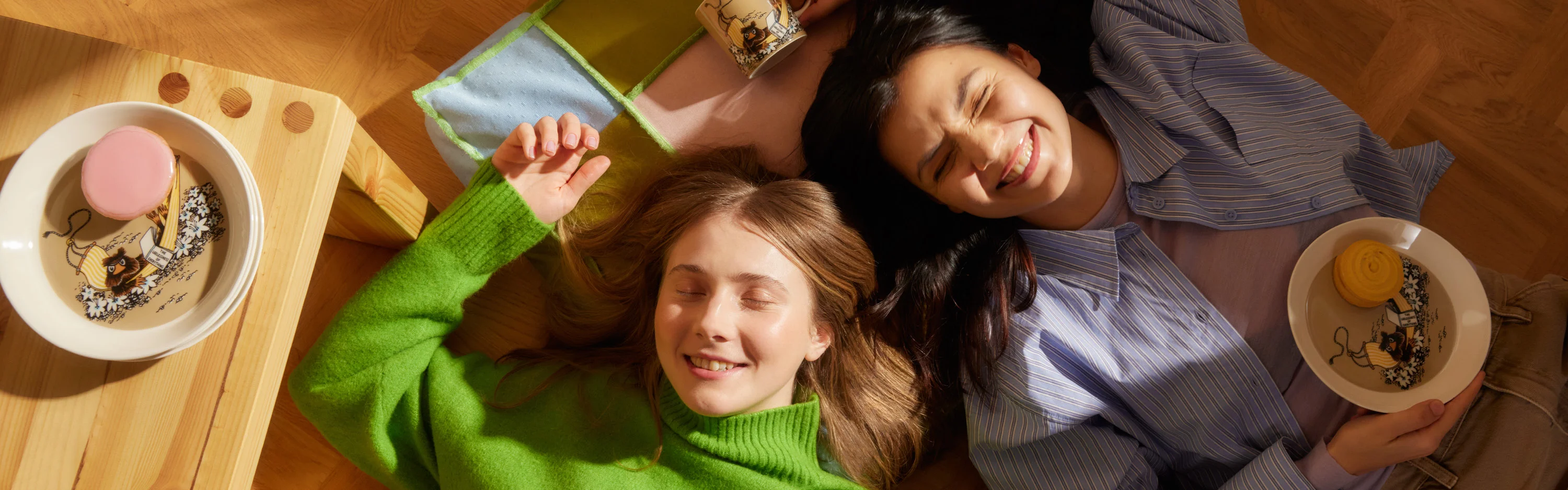Kaksi hymyilevää nuorta tyttöä makaa lattialla ympärillään Muumi-astioita.