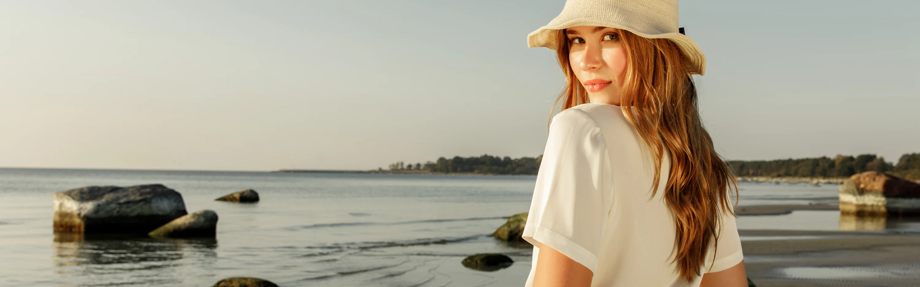 Lierihattuun pukeutunut nainen seisoo rannalla. Aurinko laskeutuu ja muuttaa kesäisen maiseman hieman utuiseksi.