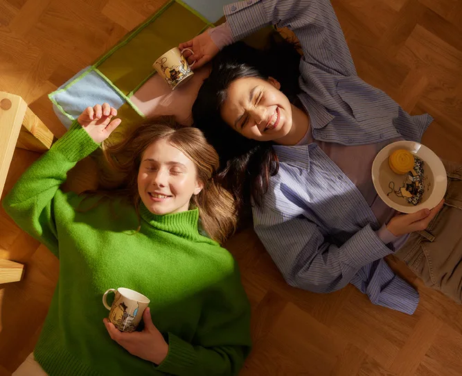 Kaksi hymyilevää nuorta tyttöä makaa lattialla ympärillään Muumi-astioita.