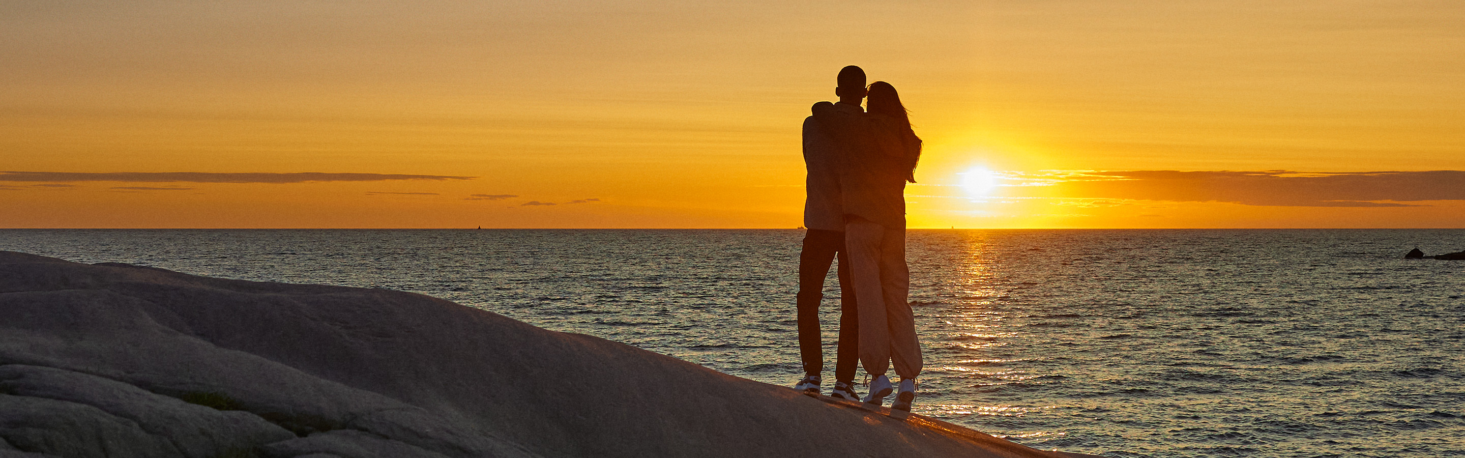 Pariskunta seisoo meren rannalla katsomassa auringonlaskua