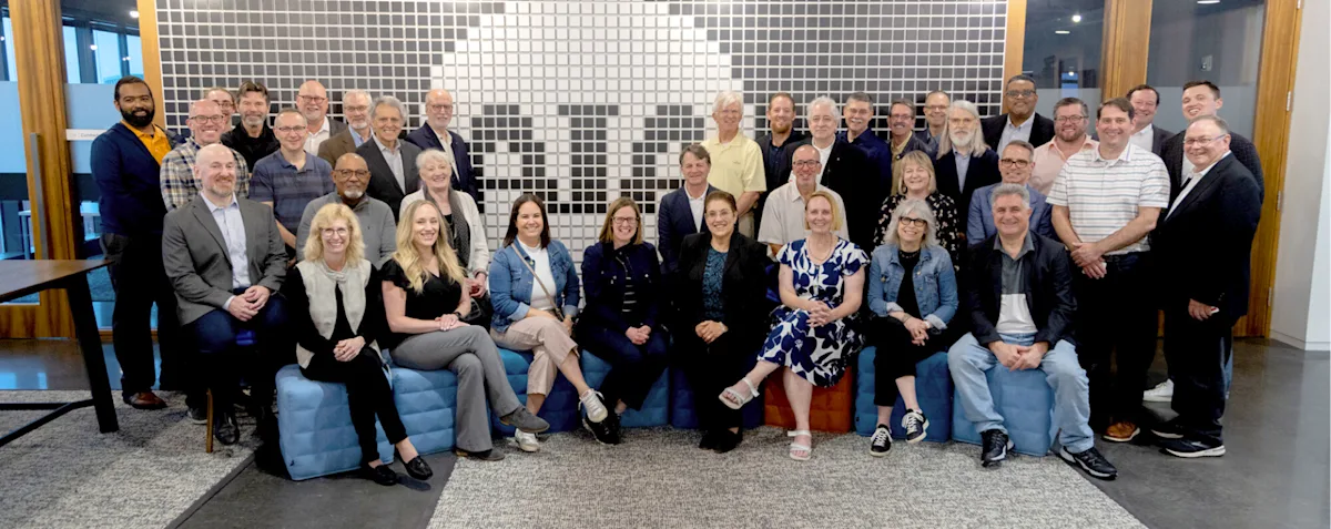 A large group photo of approximately 40 people gathered in front of a mural featuring a pattern of squares showing "AIA". The individuals, comprising both men and women, are dressed in business casual attire and are arranged in several rows, smiling for the photo.
