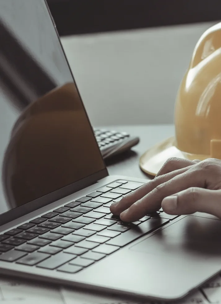 Hand on a laptop keyboard next to a yellow construction helmet on a desk.