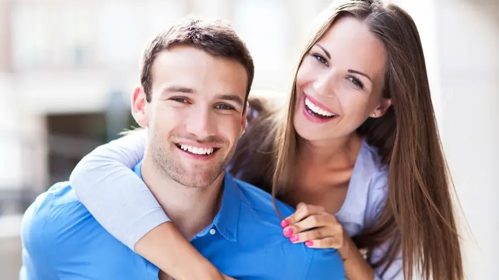 Un couple (femme et homme) sourit à la caméra, tandis que la femme enlace l’homme.